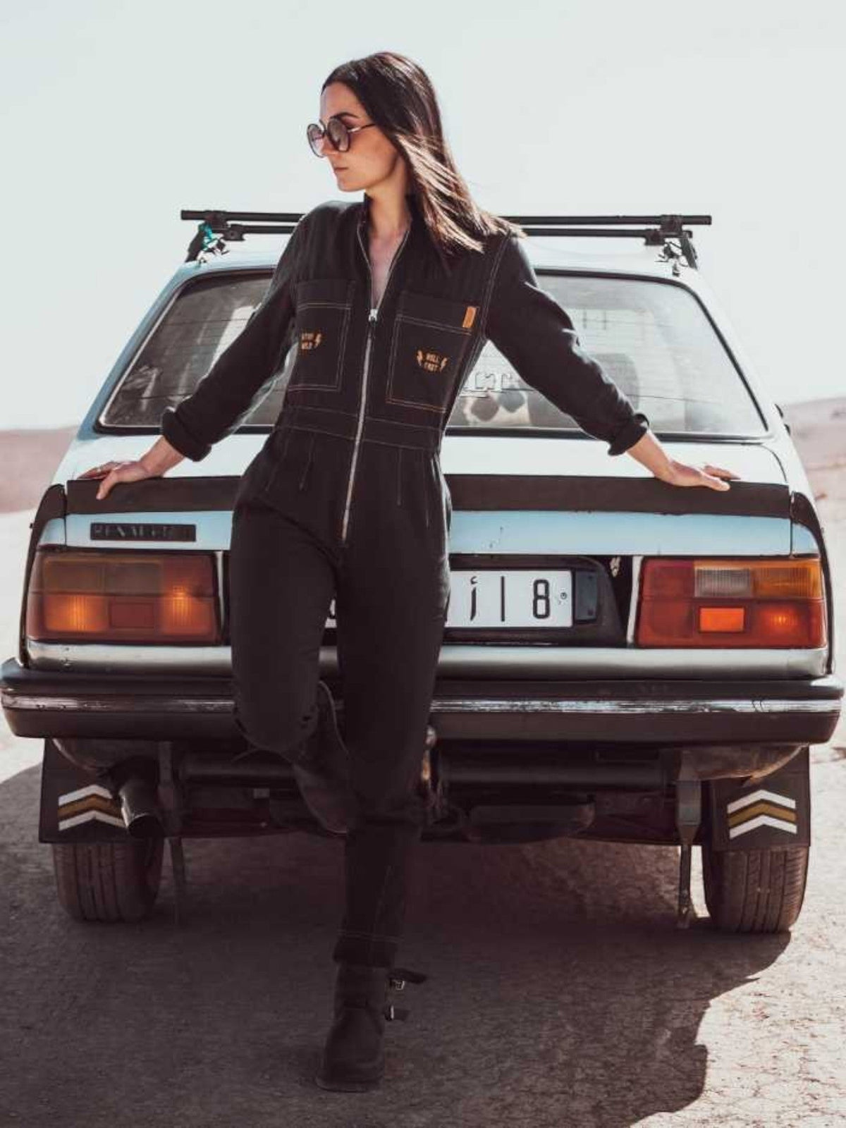 Baffle Haus Wildust black cotton aviator jumpsuit with camel stitching, worn by a woman leaning against a vintage car.