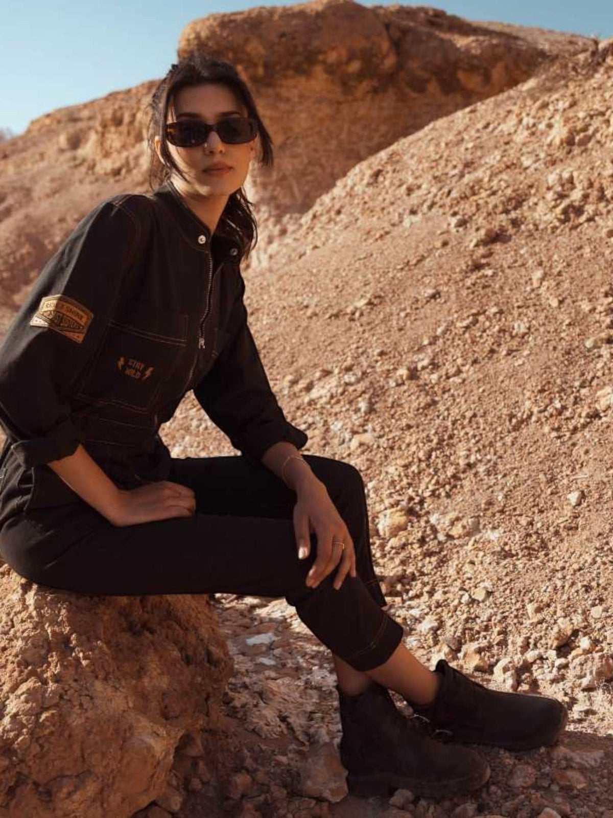Baffle Haus Wildust black cotton aviator jumpsuit with camel stitching and vintage patches, worn by a woman outdoors.