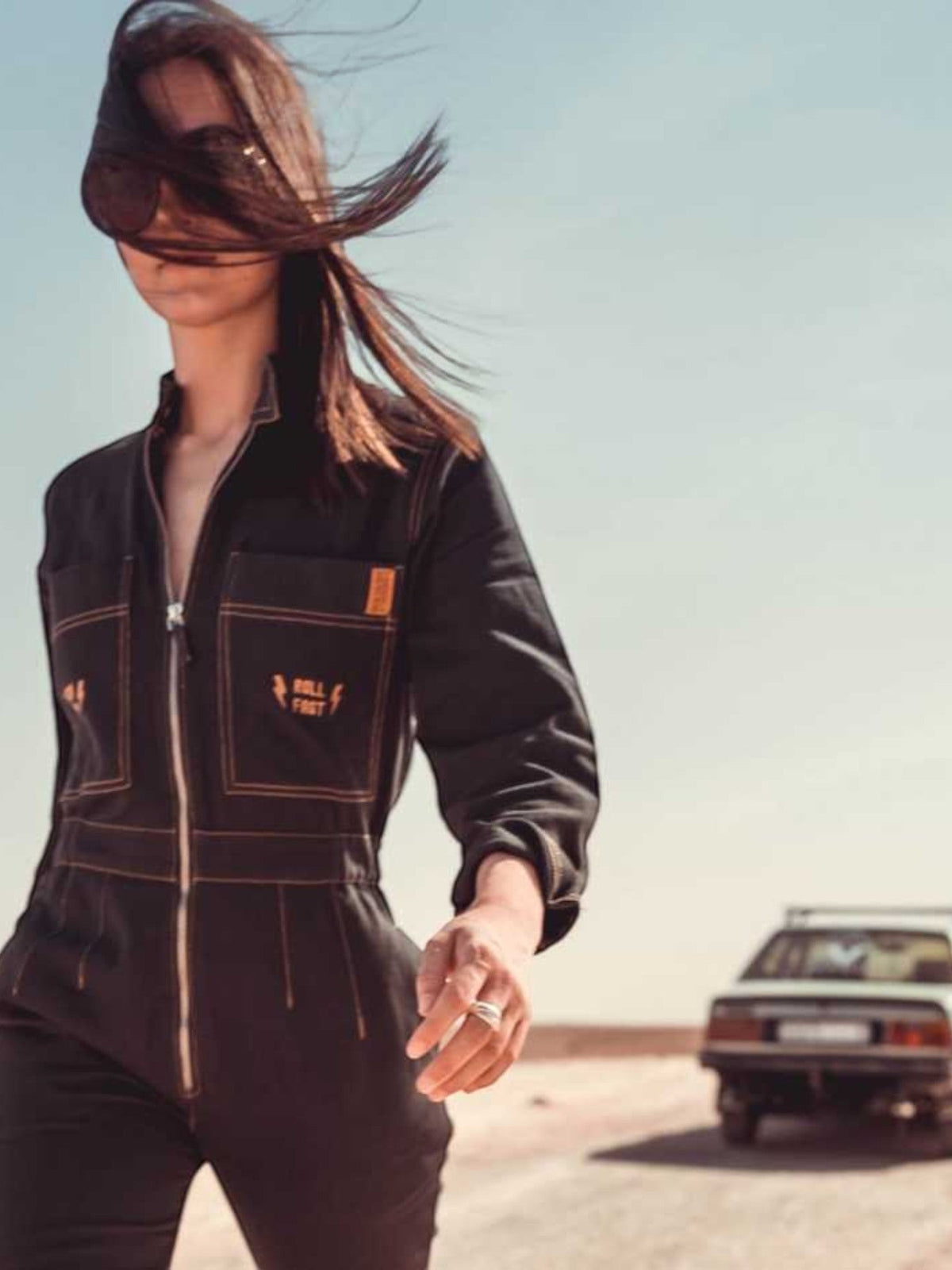 Baffle Haus Wildust black cotton aviator jumpsuit with camel stitching and chest embroidery worn by a woman outdoors