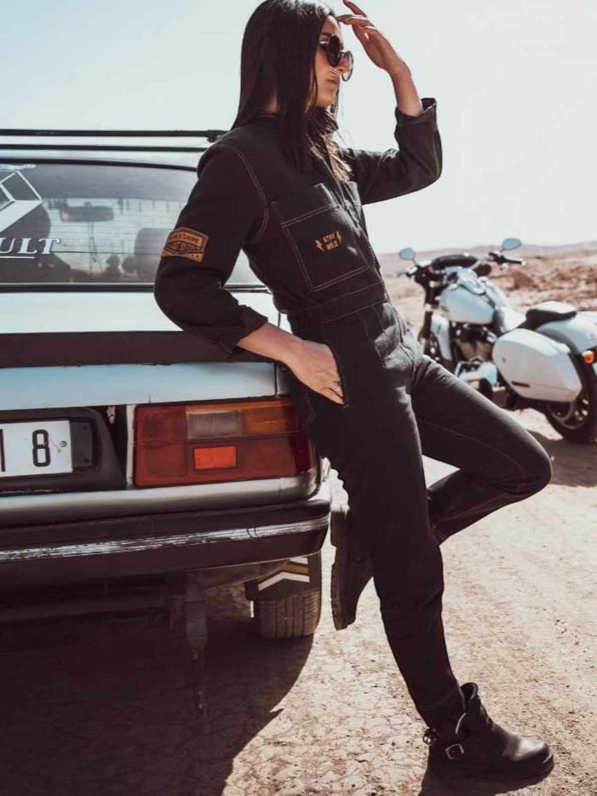 Baffle Haus Wildust black cotton aviator jumpsuit with camel stitching, worn by a woman leaning against a vintage car