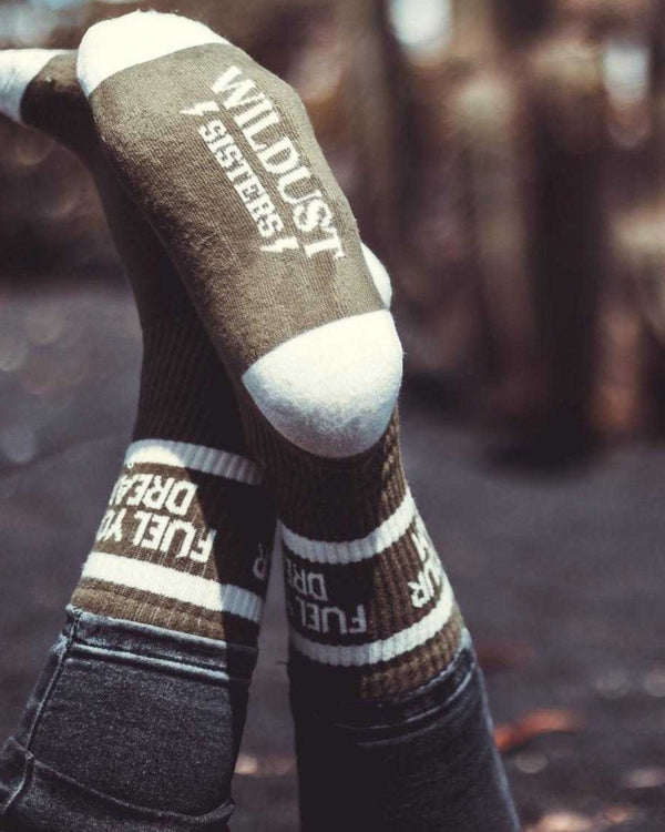 Baffle Haus Wildust Fuel Your Dreams olive green socks with white stripes and text worn on crossed legs